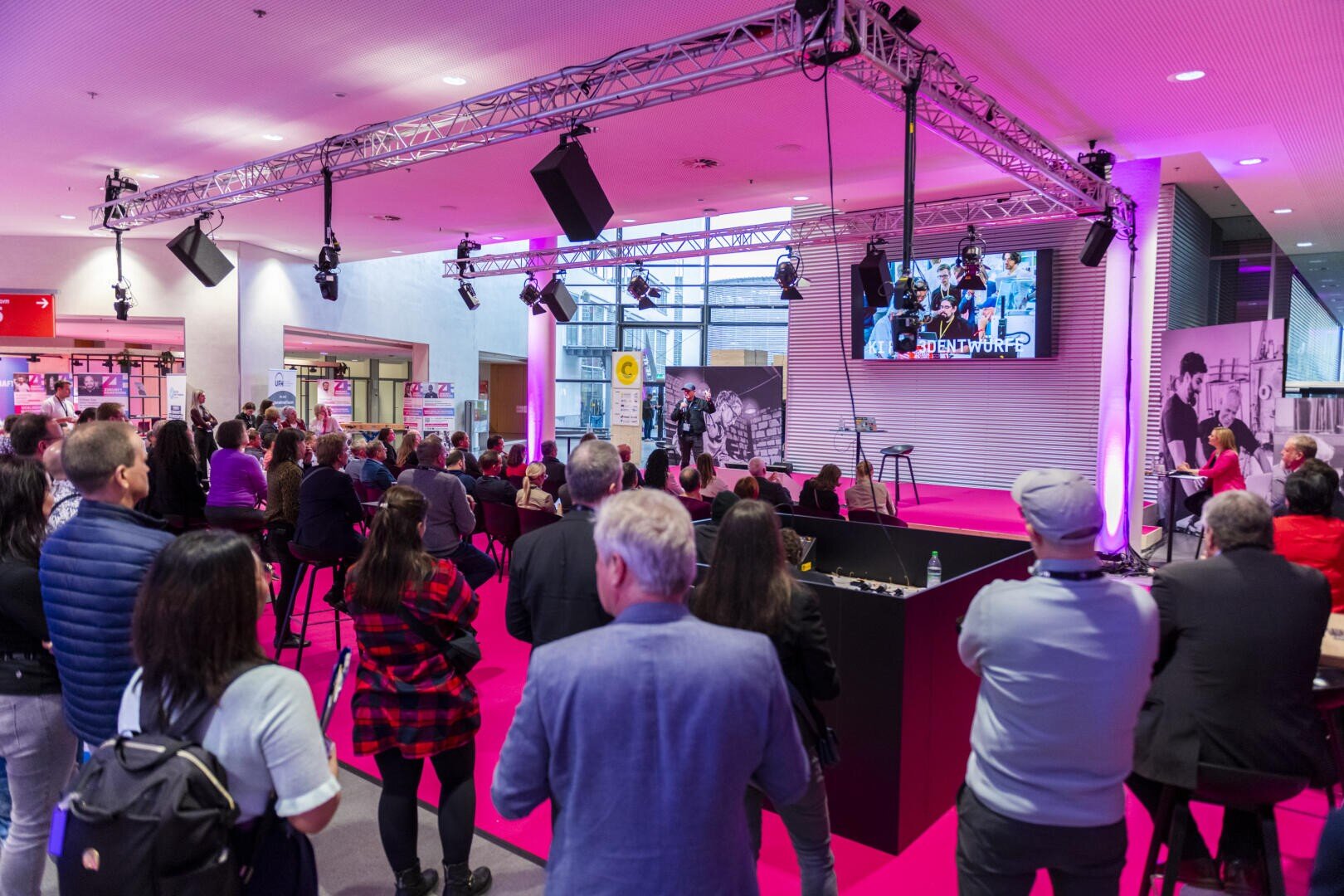 Eine Menschenmenge verfolgt einen Redner auf der Bühne in einem hell erleuchteten Veranstaltungsraum mit rosafarbenem Bodenbelag und Beleuchtung. Auf einer großen Leinwand hinter der Bühne werden Bilder gezeigt, und das Publikum ist von moderner Dekoration umgeben.