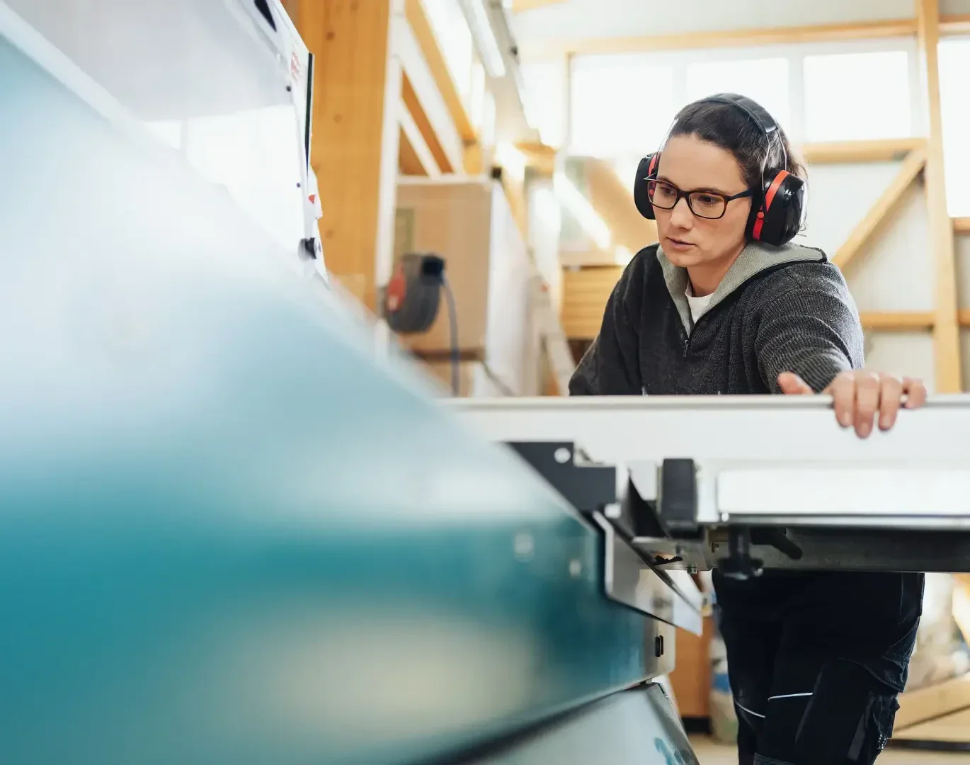 Eine Person mit Schutzbrille und Gehörschutz bedient in einer gut beleuchteten Werkstatt eine große Industriesäge und konzentriert sich darauf, das Material durch die Maschine zu führen.