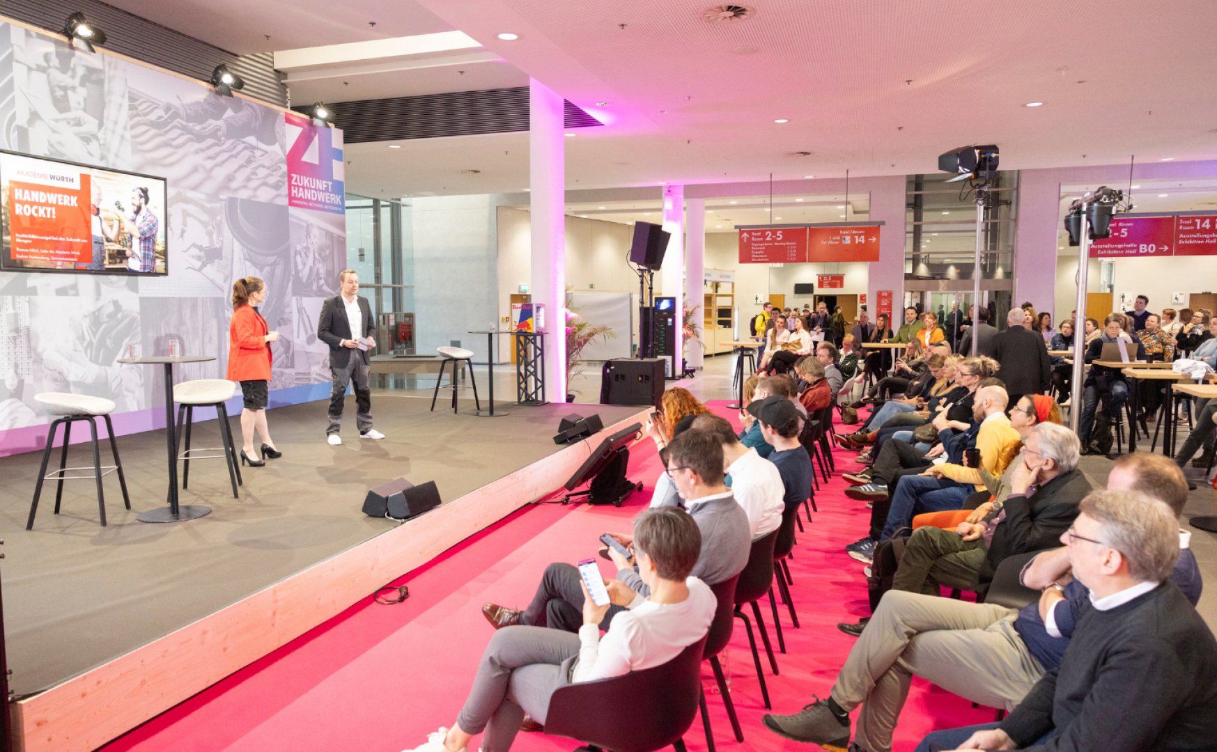 Ein Mann und eine Frau sprechen auf der Bühne zu einem sitzenden Publikum in einem hell erleuchteten Veranstaltungsraum mit rosa Teppichboden. Die Zuhörer hören aufmerksam zu, einige benutzen Telefone. Im Hintergrund sind Präsentationsbildschirme und Banner zu sehen.