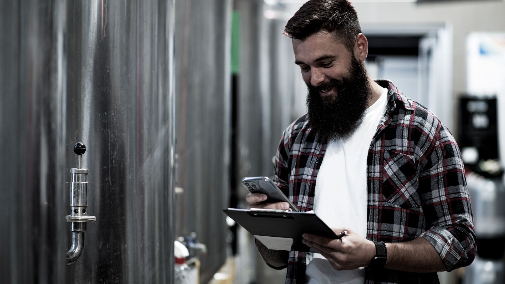Ein bärtiger Mann in einem karierten Hemd steht lächelnd in einer Brauerei, während er auf sein Telefon schaut und ein Klemmbrett in der Hand hält. Im Hintergrund sind große Edelstahltanks zu sehen.