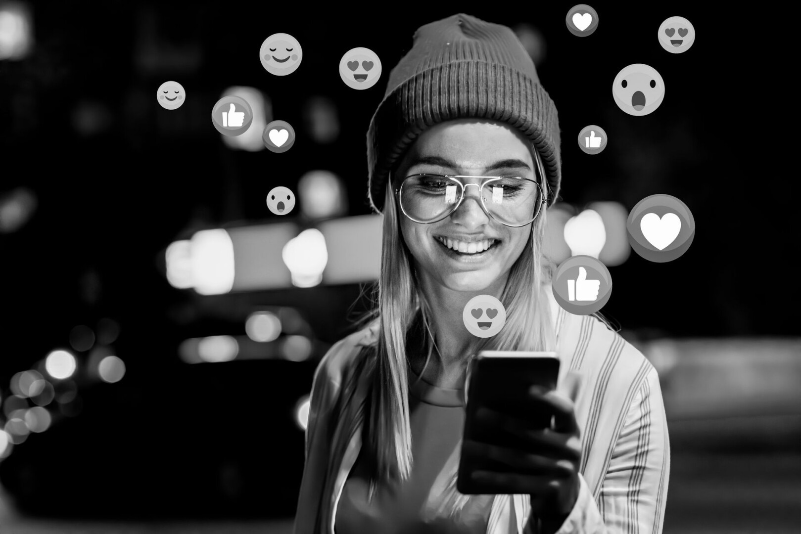 Eine lächelnde Person mit Brille und Beanie blickt auf ein Smartphone. Um sie herum schweben verschiedene Social-Media-Reaktionssymbole, wie Daumen hoch und Herzen. Der Hintergrund ist unscharf und suggeriert eine nächtliche Stadtszene.