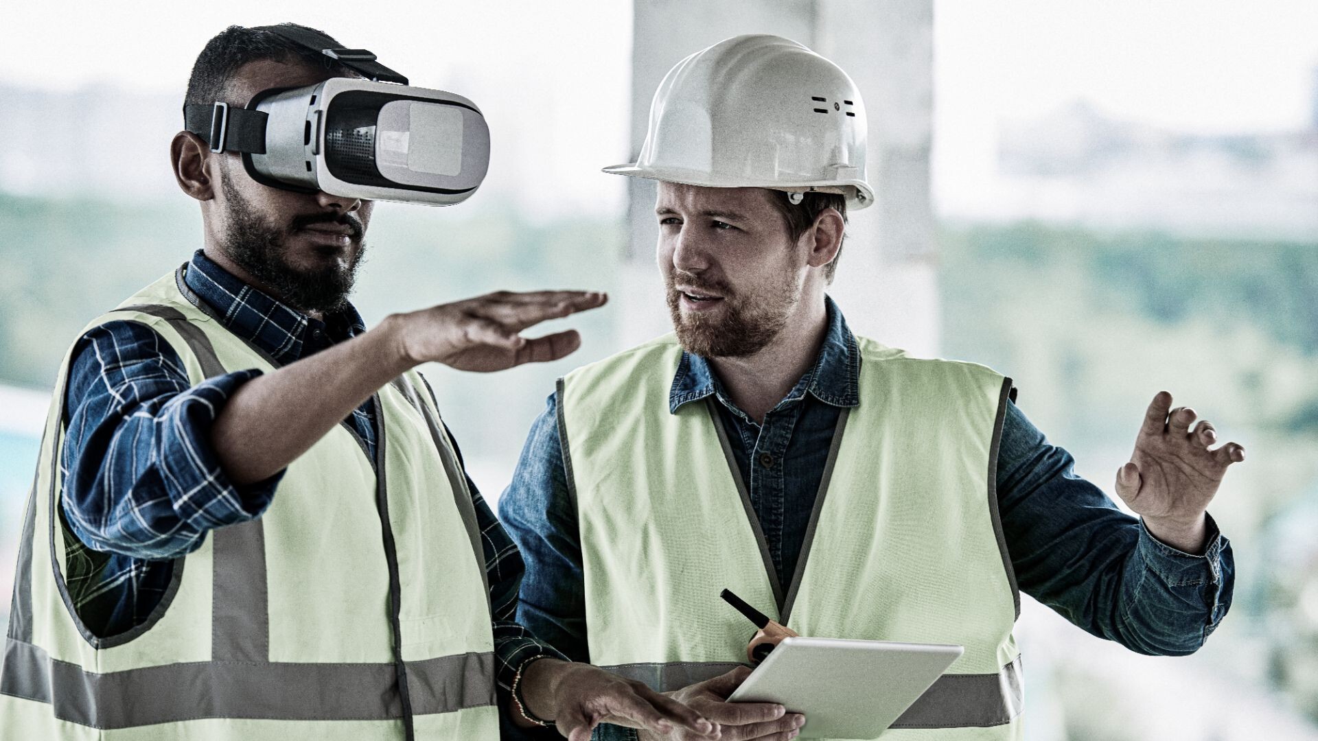 Zwei Bauarbeiter mit Sicherheitswesten und Schutzhelmen stehen im Gebäude. Einer nutzt eine Virtual-Reality-Brille und gestikuliert mit der Hand, während der andere mit Tablet und Stift zusieht und diskutiert.