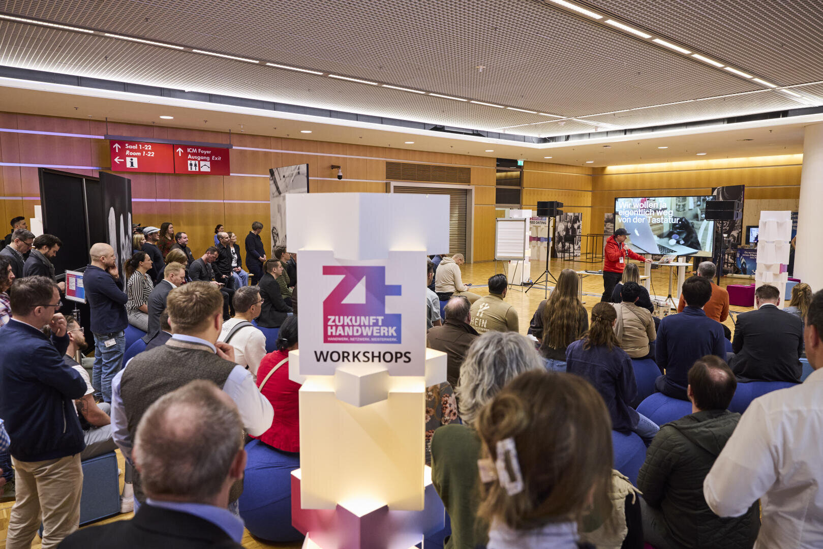 Eine Gruppe von Menschen sitzt und steht und hört einem Redner zu, der in einer modernen Halle einen Vortrag hält. Ein Schild mit der Aufschrift "Zukunft Handwerk Workshops" ist zu sehen, und auf einem großen Monitor werden vorne Dias gezeigt.