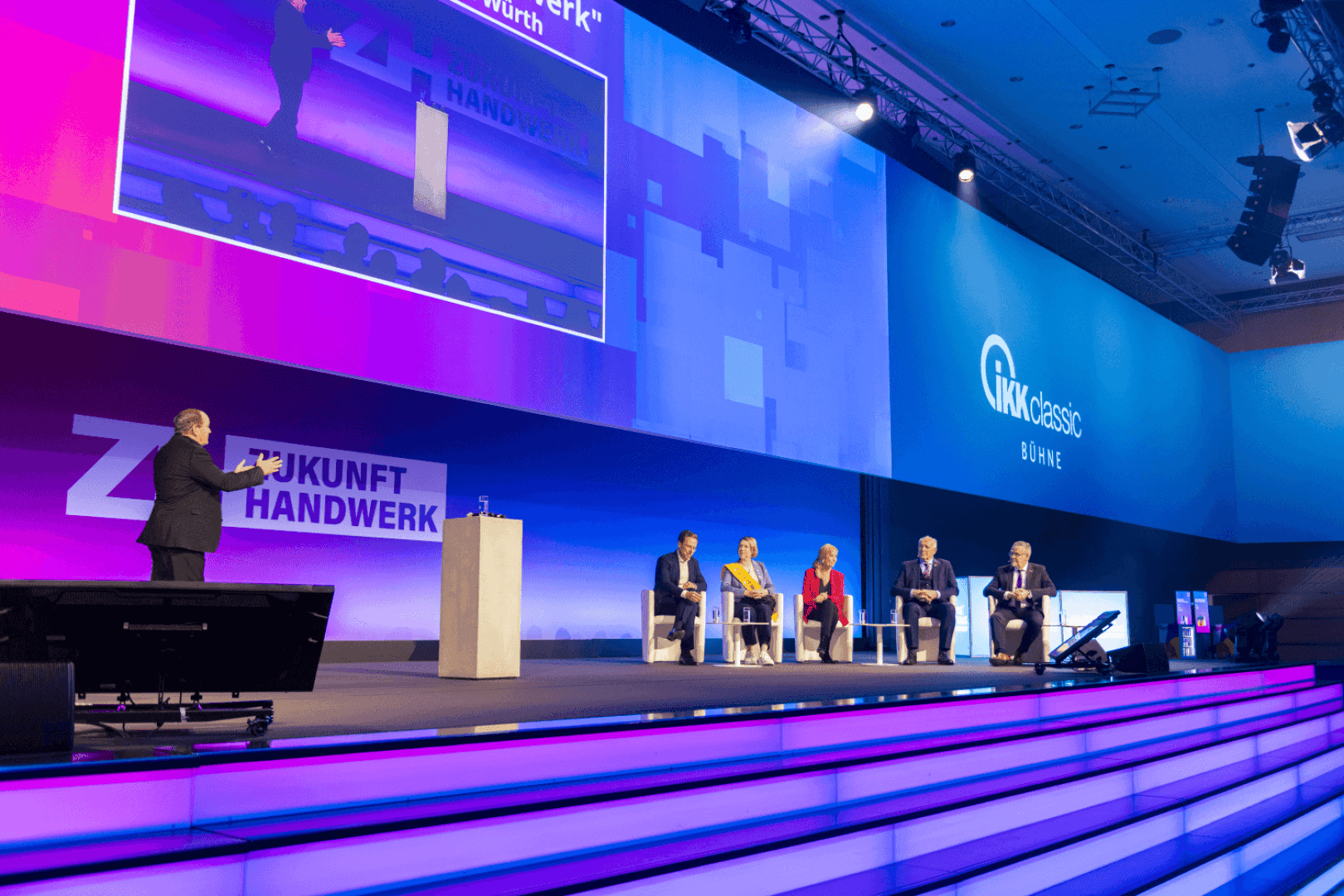 Auf einer hell erleuchteten Bühne mit großen Bildschirmen und den Logos von Zukunft Handwerk und IKK classic im Hintergrund diskutieren sechs Personen, fünf sitzend und eine stehend auf einem Podium.