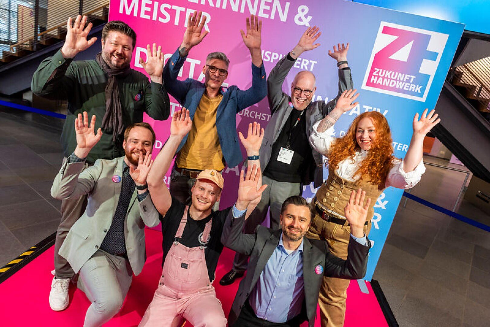 Sieben lächelnde Menschen posieren mit erhobenen Händen vor einer farbenfrohen Kulisse der Veranstaltung Zukunft Handwerk. Einige tragen Geschäftskleidung, andere tragen Arbeitsoveralls oder traditionelle Kleidung, was die Vielfalt der Berufe zeigt.