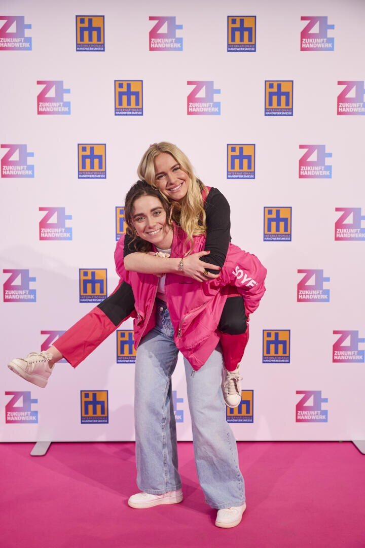 Zwei lächelnde Frauen posieren auf einem rosa Teppich; die eine in rosa Jacke und Jeans nimmt die andere, ebenfalls in Rosa, huckepack. Sie stehen vor einer Kulisse mit bunten Logos, die sich im Stechschritt wiederholen.