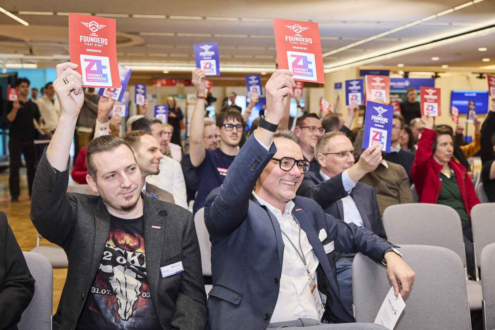 Eine Gruppe von Menschen, die bei einer Veranstaltung in einem Innenraum sitzen, lächeln und Karten mit Founders Fight Night und der Nummer 2 hochhalten. Mehrere Teilnehmer nehmen teil und schaffen eine lebhafte und engagierte Atmosphäre.