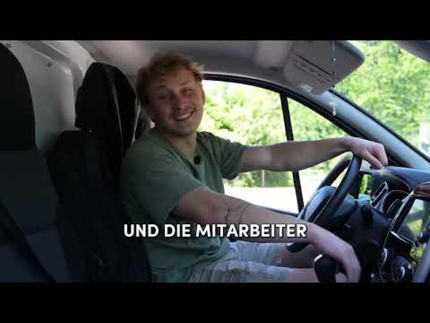 Eine lächelnde Person sitzt auf dem Fahrersitz eines Lieferwagens und blickt in die Kamera. Der deutsche Text UND DIE MITARBEITER erscheint quer über das Bild. Durch das Fenster sind Bäume und Grünflächen zu sehen.