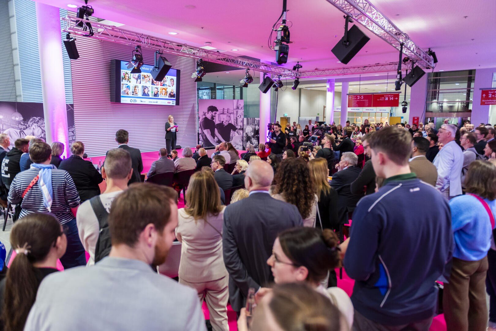 Ein großes Publikum versammelt sich in einem hell erleuchteten Veranstaltungsraum und beobachtet einen Redner, der auf der Bühne vor einer Leinwand mit Bildern spricht. Die Atmosphäre ist lebhaft und professionell, die Leute stehen und sitzen.