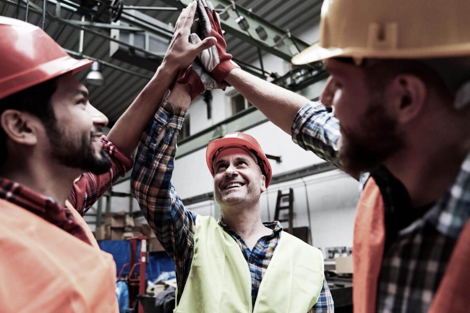 Drei Bauarbeiter, die Sicherheitswesten und Schutzhelme tragen, lächeln und klatschen sich in einem Industrielager ab.