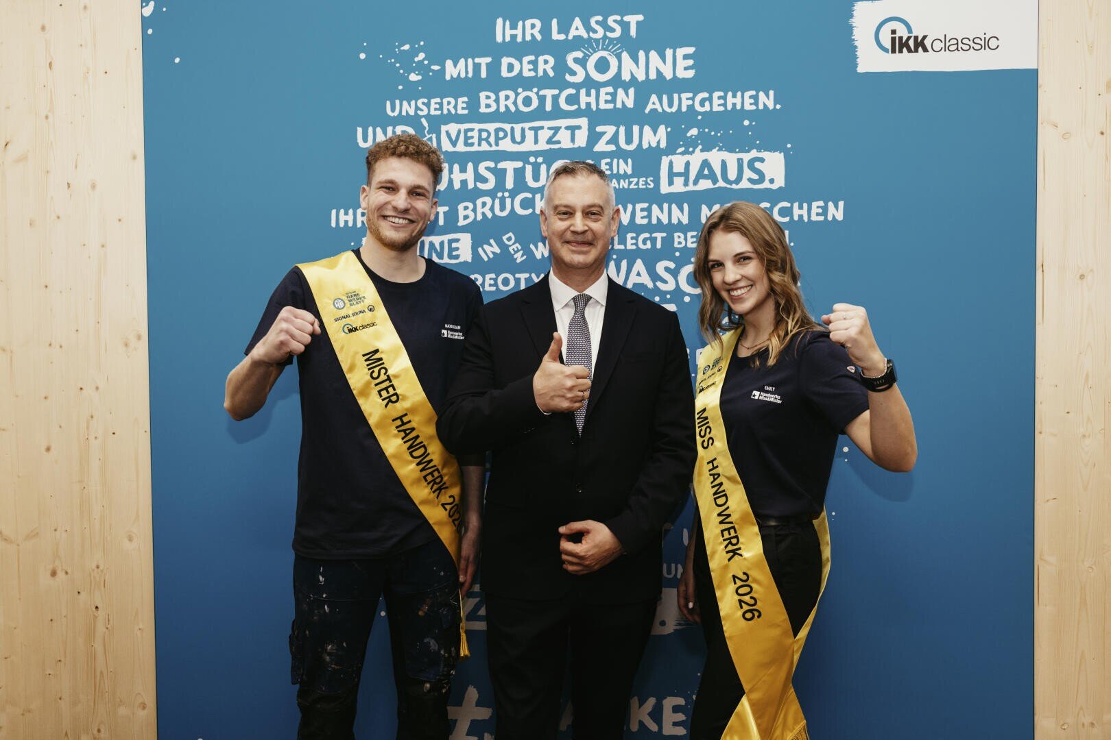 Drei Personen stehen lächelnd vor einer blauen Wand mit weißer Schrift. Die beiden an den Seiten tragen gelbe Schärpen und marineblaue Hemden, während die Person in der Mitte einen Anzug trägt und den Daumen hochstreckt. Alle wirken glücklich und feierlich.