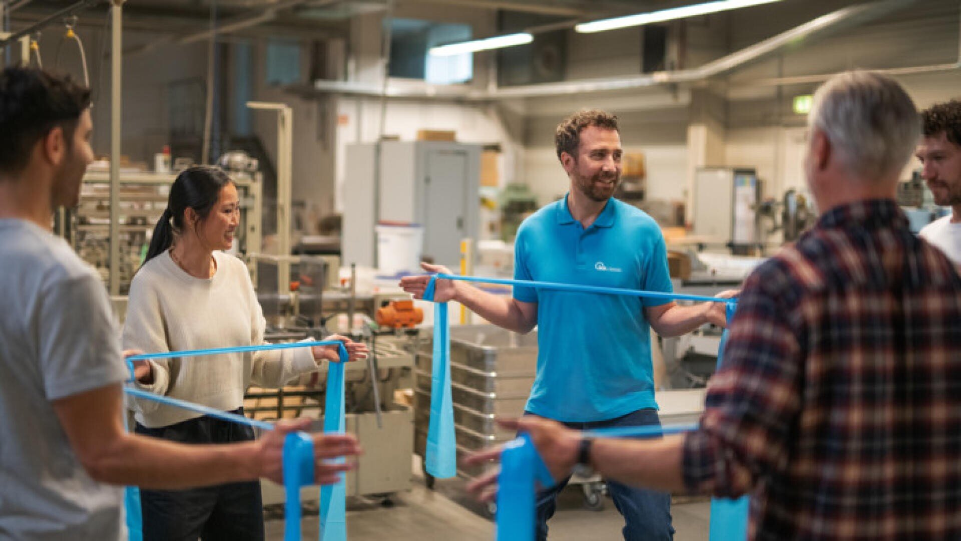 Eine Gruppe von fünf Erwachsenen steht in einem Kreis in einem Workshop, jeder hält ein blaues Gymnastikband in der Hand und lächelt, während ein Mann in einem blauen Poloshirt die Aktivität anleitet.